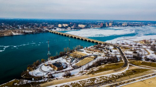 Aerial view of a river with ice, a bridge, and a city skyline in the background, surrounded by snow-covered land.