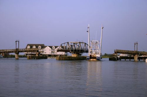 A drawbridge over calm water with a fishing boat nearby and a white building in the background under a clear sky.