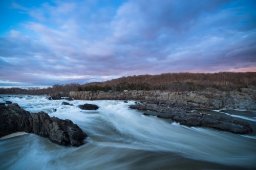 A flowing river cascades over rocky terrain under a cloudy sky at dusk, with trees lining the distant shore.