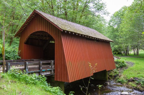 A red covered bridge spans a small stream, surrounded by lush green trees and grass.