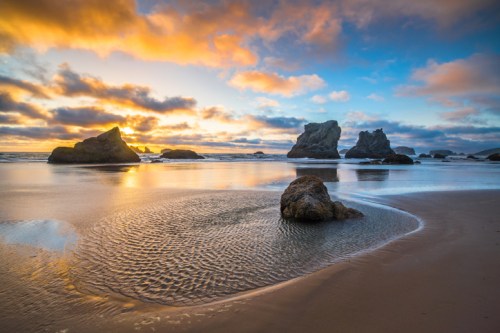 A serene beach at sunset, featuring rocky formations and gentle waves reflecting vibrant orange and blue skies.