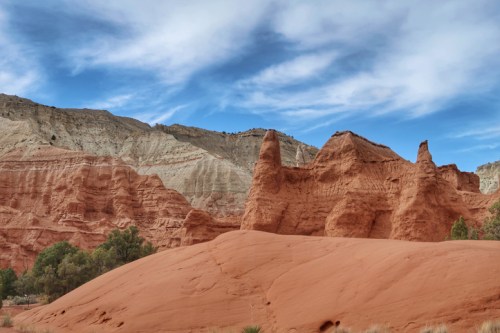 Red rock formations rise against a blue sky with wispy clouds, surrounded by green vegetation in a desert landscape.