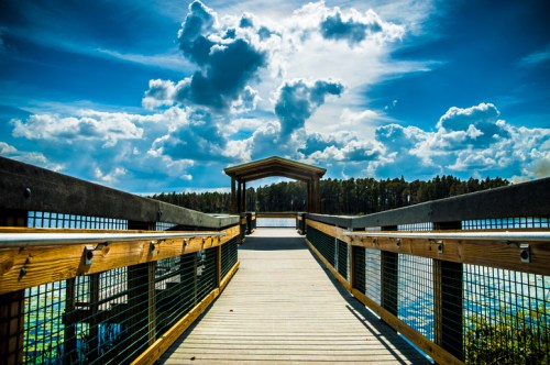 A wooden boardwalk leads to a gazebo over a serene lake, under a bright blue sky with fluffy clouds.