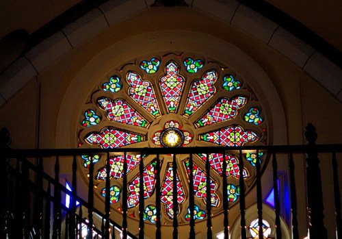 A colorful stained glass rose window with intricate patterns, illuminated by light, set in a church interior.
