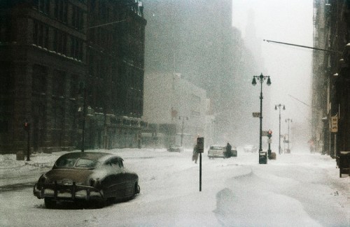 A snowy street scene with vintage cars buried in snow, buildings shrouded in fog, and streetlights barely visible.