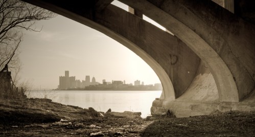 A view of a city skyline through the arch of a bridge, with calm water and a muted, sepia-toned atmosphere.