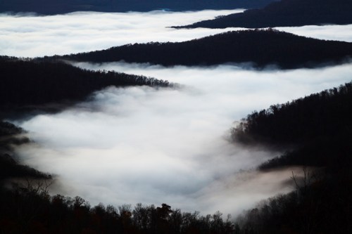 A misty valley with rolling fog covering the landscape, surrounded by dark, tree-covered hills.