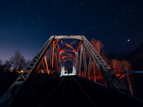 A railway bridge illuminated at night, surrounded by trees and a starry sky, creating a serene, atmospheric scene.