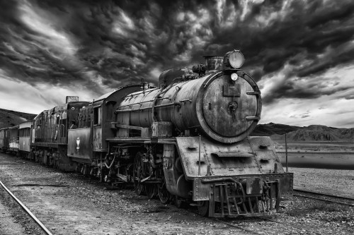 A vintage steam locomotive sits on tracks under a dramatic, cloudy sky in black and white.