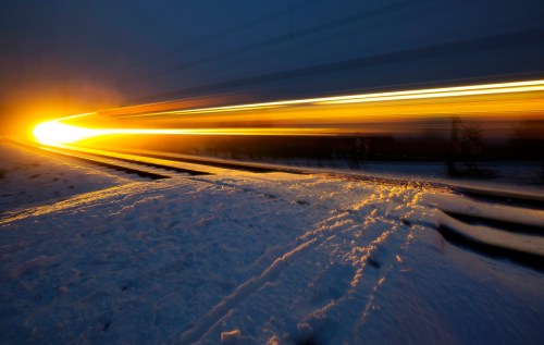 A train moves through a snowy landscape at night, leaving a trail of bright light against the dark sky.
