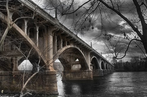 A historic stone bridge arches over a river, surrounded by bare trees and a dramatic cloudy sky.