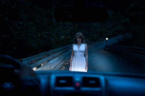 A woman in a white dress stands on a dark road, illuminated by headlights, with trees in the background.