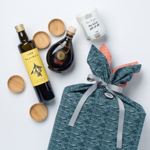 A gift set featuring olive oil, balsamic vinegar, a candle, and wooden coasters, all wrapped in a patterned fabric.