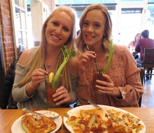 Two smiling women hold drinks with celery, sitting at a table with food in a cozy restaurant setting.