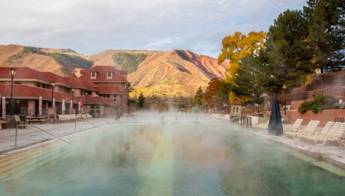 A steaming outdoor pool surrounded by mountains and autumn foliage, with lounge chairs and buildings in the background.