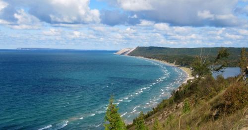 A scenic view of a coastline with rolling waves, sandy beach, and lush green hills under a partly cloudy sky.