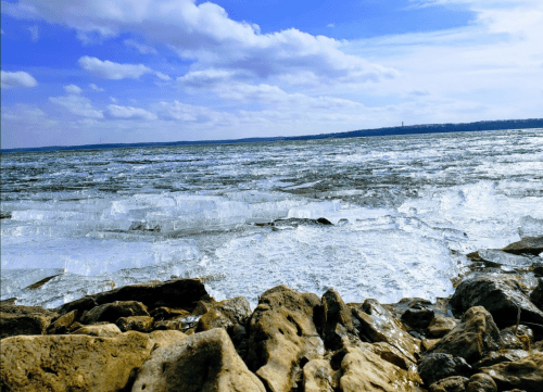A rocky shoreline with ice formations on a lake under a blue sky with fluffy clouds.