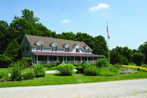 A large, two-story house with a porch, surrounded by greenery and flowers, under a clear blue sky. An American flag is visible.