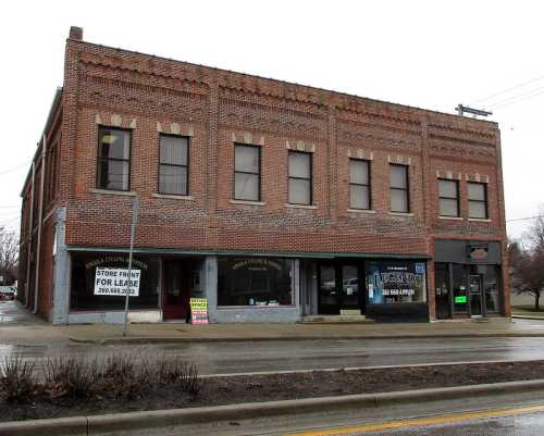 A brick building with multiple windows and storefronts, featuring a "For Lease" sign, on a rainy day.