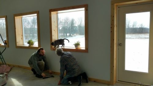 Two people work on a window ledge while a dog walks along the ledge, with a snowy landscape visible outside.