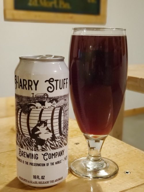 A can of Harry Stuff Brewing Company beer next to a filled glass, showcasing a deep red beverage on a wooden table.