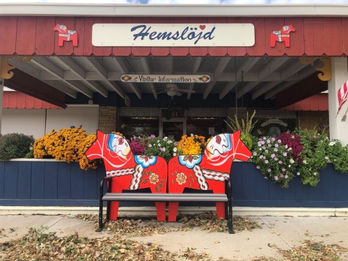 Colorful wooden horse sculptures sit on a bench in front of a visitor information center, surrounded by vibrant flowers.