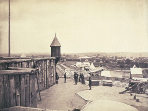 Historic black-and-white photo of a fort with soldiers, wooden structures, and a distant landscape.