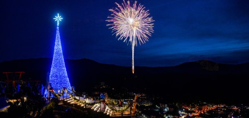 A brightly lit blue Christmas tree with fireworks in the night sky above a scenic mountain town.