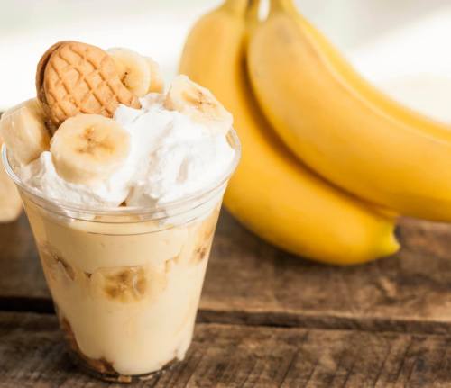 A clear cup filled with banana pudding, topped with whipped cream and banana slices, next to ripe bananas on a wooden surface.