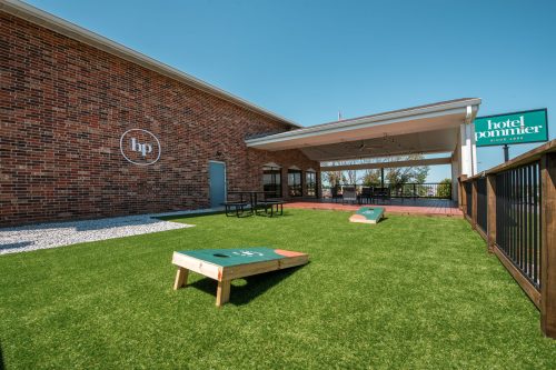 Outdoor area of Hotel Pommier featuring cornhole boards, green turf, and a brick building under a clear blue sky.