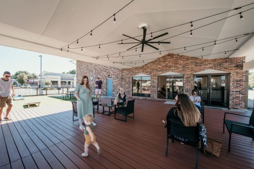 A spacious outdoor area with people socializing, sitting on chairs, and a child playing on a wooden deck.