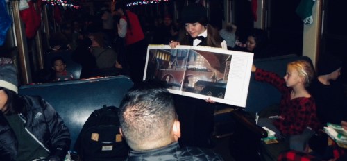 A person in a tuxedo holds a large poster while seated passengers look on in a festive train interior.