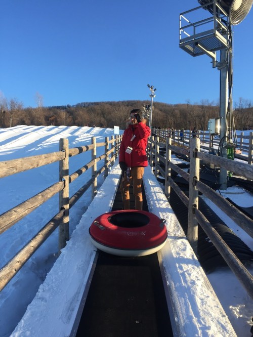 A person in a red jacket stands on a snowy slope next to a snow tubing conveyor belt.