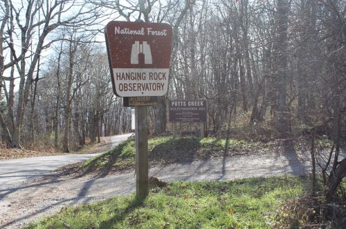 Sign for Hanging Rock Observatory and Potts Creek in a wooded area, with a gravel road and sunlight filtering through trees.