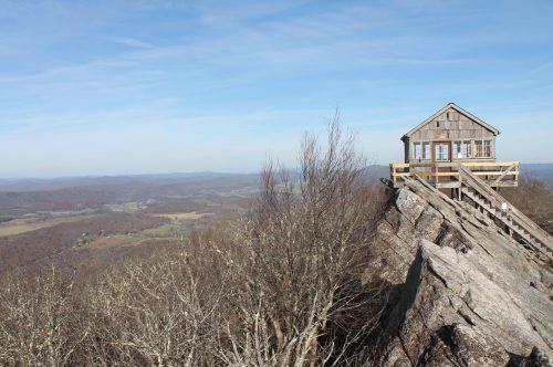A wooden cabin perched on a rocky outcrop, overlooking a vast valley and distant mountains under a clear blue sky.