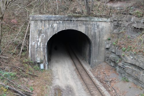 A dark tunnel entrance surrounded by rocky terrain and trees, with railway tracks leading inside.