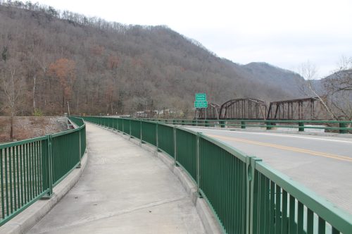A paved walkway alongside a river, leading to a bridge with mountains in the background and a sign nearby.