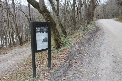 A sign along a dirt road in a wooded area, with trees and a river visible in the background.