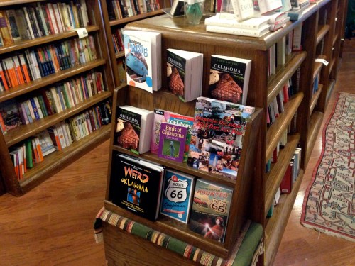 A wooden bookshelf displays various books about Oklahoma, including travel guides and local history.
