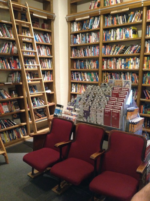 A cozy library with wooden shelves filled with books, a ladder, and three red chairs in front.