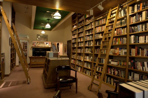 A cozy library with tall bookshelves, wooden ladders, and a central checkout desk, illuminated by warm lighting.