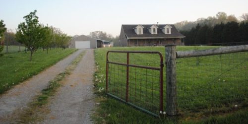 A gravel driveway leads to a house and barn, surrounded by green fields and trees under a clear sky.