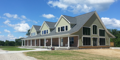 A large, modern house with a green exterior, multiple gables, and large windows, set against a blue sky with clouds.
