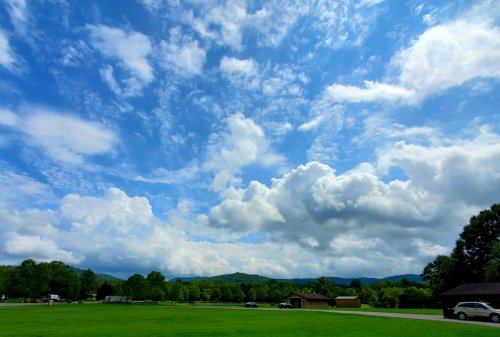 A vibrant sky filled with fluffy clouds over a green landscape and distant hills, with a few buildings and cars visible.