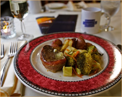 A plate of meatloaf with mashed potatoes, broccoli, and carrots, set on a decorative table with utensils and a drink.