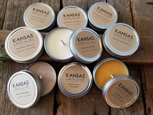 A collection of Kansas Earth and Sky candles in metal tins, featuring various scents, arranged on a wooden surface.