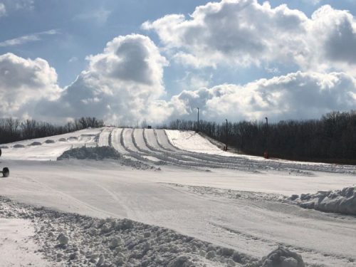 A snowy landscape with a hill and ski tracks under a bright sky filled with fluffy clouds.