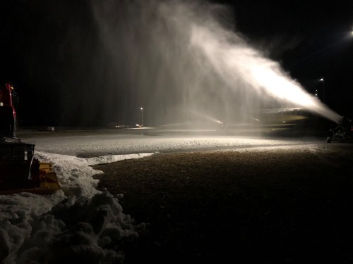 Snowmaking equipment operates at night, spraying artificial snow over a dark landscape with lights illuminating the area.