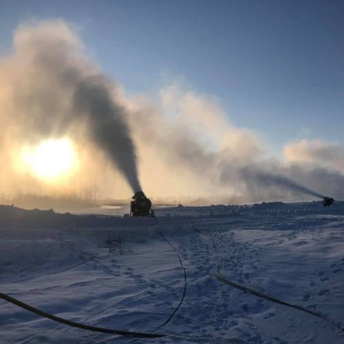 Two machines emit smoke in a snowy landscape, with a bright sun rising in the background and footprints in the snow.