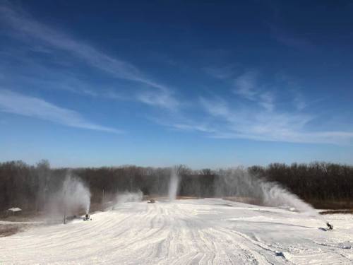 Snowmaking machines create artificial snow on a winter landscape under a clear blue sky.